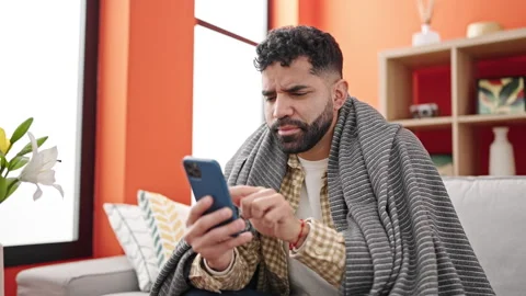 Young hispanic man sitting on sofa covering with blanket talking on smartph.. Video stock 245047261