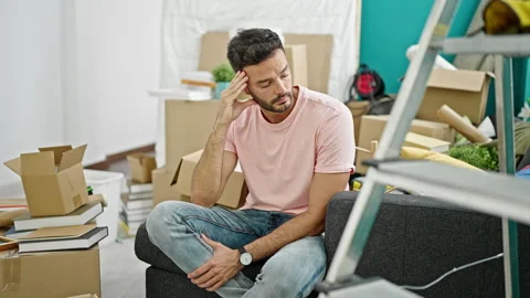 Young hispanic man sitting on sofa with serious expression at new home Stock Footage 247378845