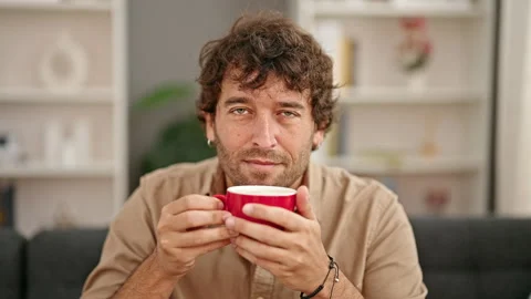 Young hispanic man smelling cup of coffee sitting on sofa smiling at home Video stock 245045326