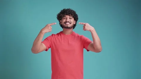 Young hispanic man smiling and pointing at his teeth and beautiful smile on blue Video stock 239360892