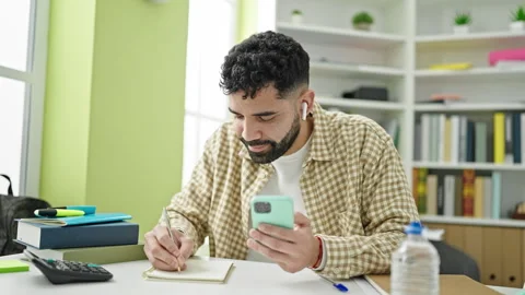 Young hispanic man student using smartphone and earphones writing notes at .. Stock Footage 245046848