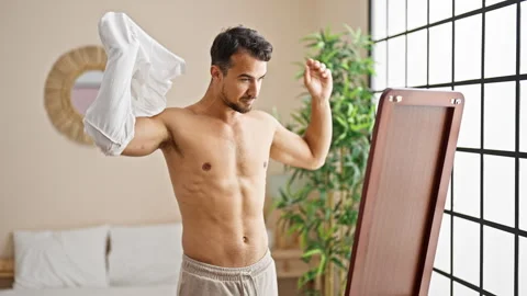 Young hispanic man taking t shirt off looking body on mirror at bedroom Stock Footage 249911929