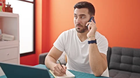 Young hispanic man talking on smartphone using laptop taking notes at dinni.. Video stock 243489750