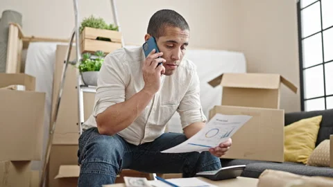 Young hispanic man talking on smartphone reading document at new home Stock Footage 245023098