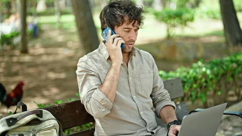 Young hispanic man talking on smartphone using laptop sitting on bench at par Stock Footage 245559536