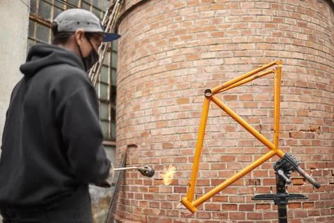 Young Hispanic man using a blowtorch to remove the paint of a bicycle frame Foto stock