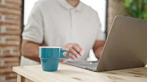 Young hispanic man using laptop drinking coffee at dinning room Stock Footage 245023435
