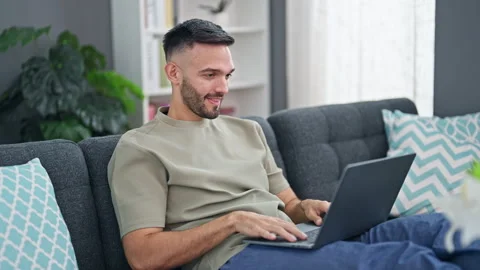 Young hispanic man using laptop sitting on sofa at home Stock Footage 247382429