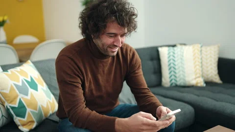 Young hispanic man using smartphone sitting on sofa at home Video stock 233403160