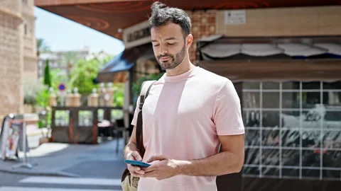 Young hispanic man using smartphone wearing backpack at coffee shop terrace Video stock 245037492