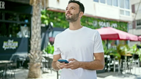 Young hispanic man using smartphone smiling at coffee shop terrace Video stock 245037642