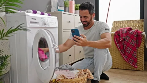 Young hispanic man using smartphone washing clothes smiling at laundry room Video stock 245080461