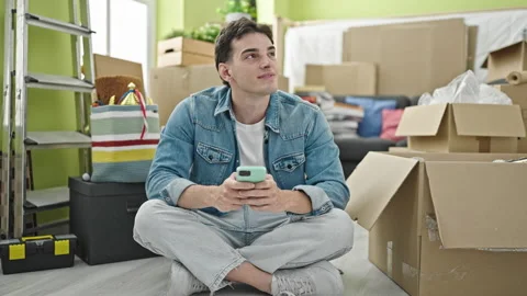Young hispanic man using smartphone sitting on floor at new home Stock Footage 246123461