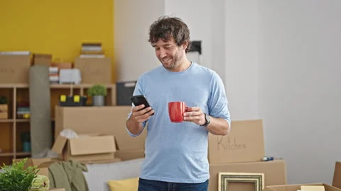 Young hispanic man using smartphone drinking coffee standing at new home Video stock 246132276