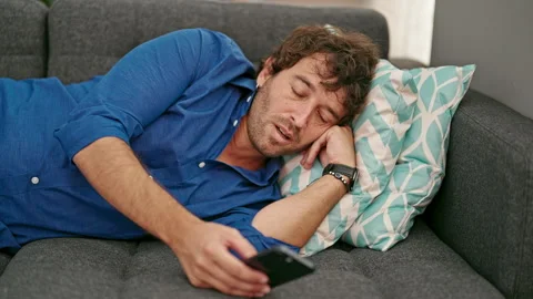 Young hispanic man using smartphone lying on sofa tired at home Video stock 247379208