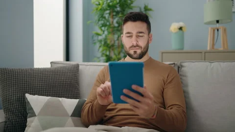 Young hispanic man using touchpad sitting on sofa at home Stock Footage 173780494