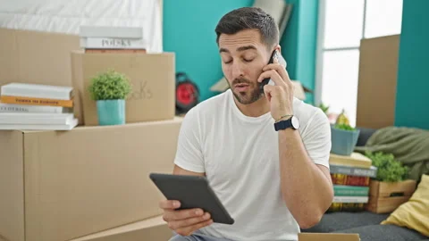 Young hispanic man using touchpad talking on smartphone at new home Video stock 243489898