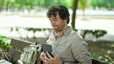Young hispanic man using touchpad sitting on bench smiling at park Video stock 245046028