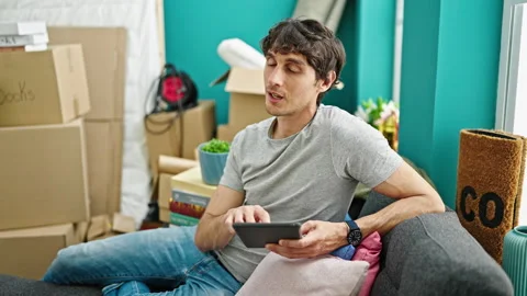 Young hispanic man using touchpad sitting on sofa at new home Video stock 247384968