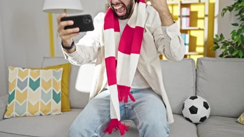 Young hispanic man watching soccer match sitting on sofa celebrating at home Stock Footage 247378342