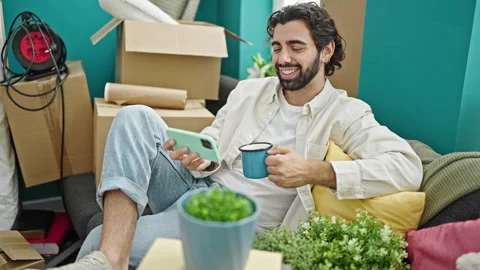 Young hispanic man watching video on smartphone drinking coffee at new home Stock Footage 247378406