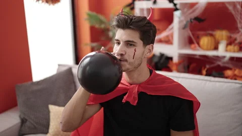 Young hispanic man wearing devil costume inflating balloon at home Stock Footage 236566923