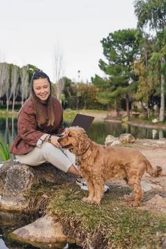 A young Hispanic woman with long dark hair sits on a rock by a pond, smiling at Stock Photos
