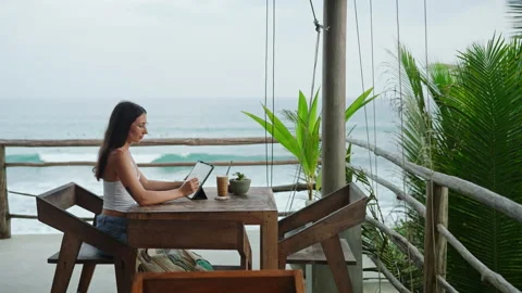 Young illustrator drawing on tablet at cafe with ocean view, palm trees. Pretty Stock Footage 253822066