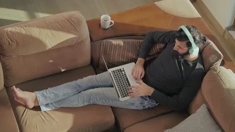 Young Indian bearded man lying on couch with headphones on &amp; working on a Laptop Stock Footage 165341931