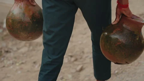 A young Indian boy in flip flops carries a two clay jug of water in his hands Video stock 303869948