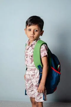A young Indian boy stands with a backpack, his expression serious, against a  Stock Photos