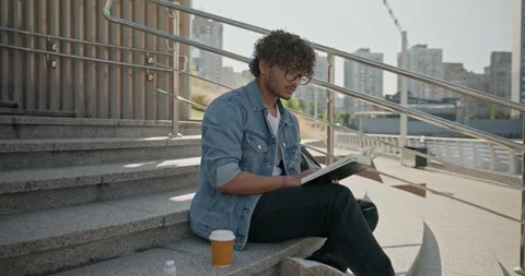 Young indian guy student reading book sitting on a stairs outdoor Stock Footage 246790973