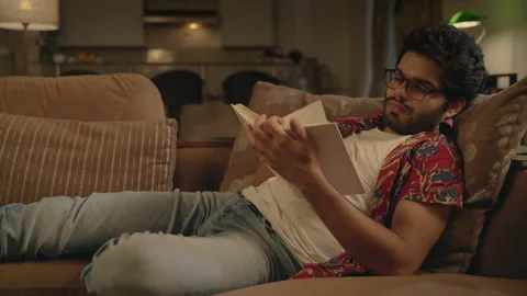 Young Indian man lying on a couch comfortably reading a book at home in night. Stock Footage 170572915