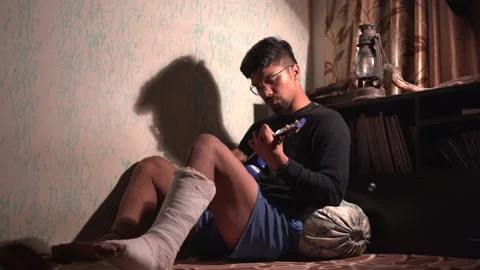 Young Indian man playing ukelele while sitting on the bed with a fractured le Stock Footage 147237527