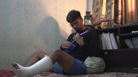 Young Indian man playing ukelele while sitting on the bed with a fractured le Stock Footage 147237545