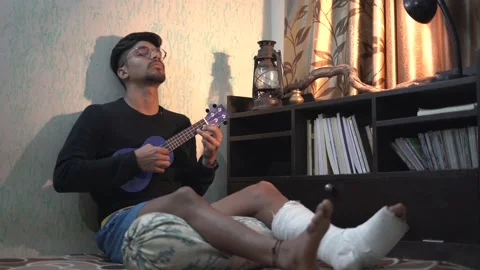 Young Indian man playing ukelele while sitting on the bed with a fractured le Stock Footage 147237596