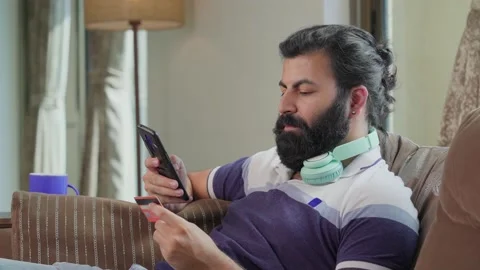 Young Indian man sitting on couch making online transaction through Mobile Phone Stock Footage 165410137