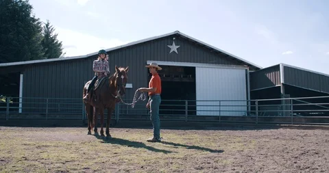 Young instructor teaching female student on horseback near western barn Stock Footage 98315720