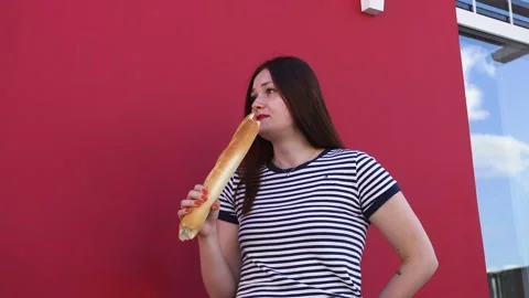 A young lady bites off a piece of bread, a baguette against a red wall, laughs Stock Footage 137537408