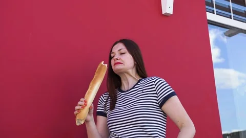A young lady bites off a piece of bread, a baguette against a red wall, laughs Stock Footage 137537453