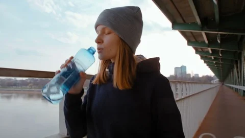 Young Lady is Drinking Water from Bottle on the Bridge over the River and City Vidéo 74250161