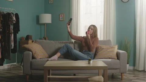 Young lady with red apple sits on couch and takes selfie on cell phone 스톡 동영상 149965383