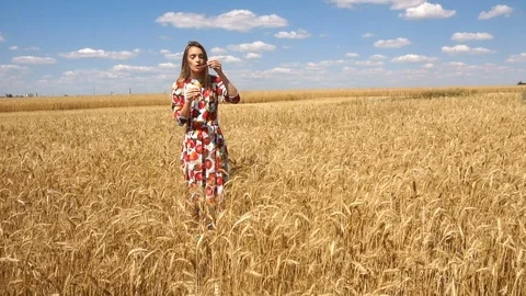 Young lady standing in a wheat field against blue sky and inflates soap-bubbles Stock Footage