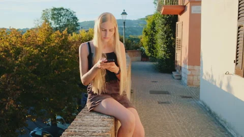 Young lady using smartphone sitting on the street in sun rays Stock Footage 143013763