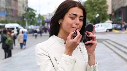 Young lady while looking on the mirror make her lips red lipstick on the street. Stock Footage 75582660