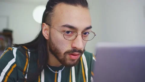Young latin long-haired man in shock as he looks at his computer, wearing gla Stock Footage 153334949