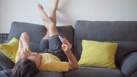 Young Latin man lying on sofa with head down and legs up texting on cell ph.. Stock Footage 238875258