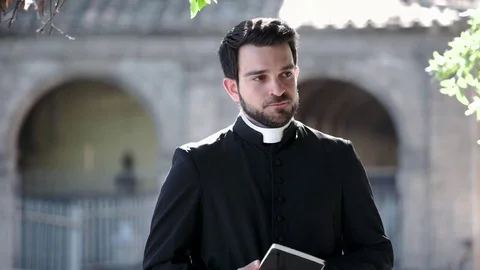 Young latin priest scrolls the gospels and smiles at the camera- outdoors Vidéo 76434963
