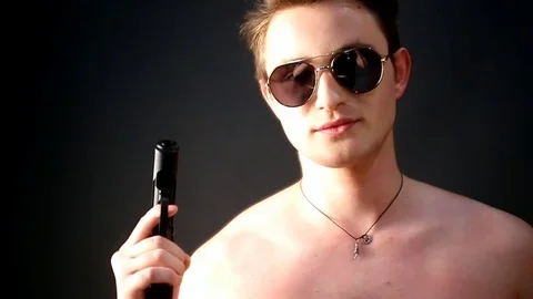 A young lean white guy with a naked torso in sunglasses holds in his hand a gun Stock Footage 83247178