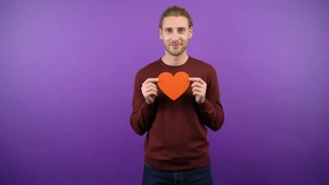 Young long-haired guy with big paper red heart on isolated purple background 4k Stock Footage 148278459